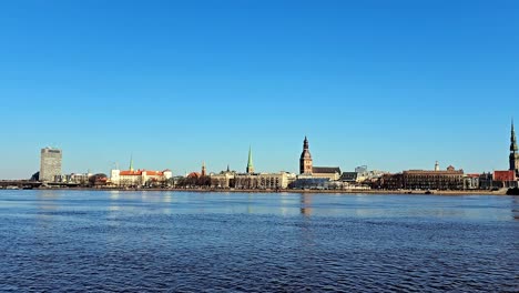 Eine-Weite-Aufnahme-Der-Skyline-Einer-Stadt-über-Einem-Ruhigen-Fluss-Daugava,-Mit-Einer-Stadtbrücke-Von-Riga-Im-Vordergrund