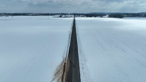 Drone-flyover-empty-road-surrounded-by-snow-covered-fields-in-scenic-area-of-America