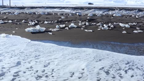 Tomas-Cinematográficas-Aéreas-Capturadas-Por-Un-Dron-De-4k-Muestran-Olas-Rompiendo-Que-Llegan-A-La-Costa,-Con-Hielo-Roto-De-Los-Glaciares-Esparcidos-Por-El-Suelo