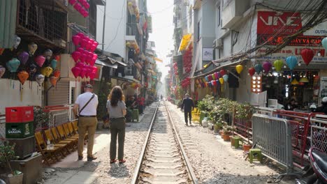 Paar-Erkundet-Die-Lebhafte-Zugstraße-In-Hanoi-Mit-Geschäften-Und-Bunten-Laternen,-Sonniger-Tag