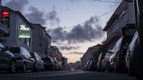 Los-Coches-Que-Viajan-Pasan-Junto-A-Los-Coches-Estacionados-En-Una-Calle-Con-Hoteles-Y-Tiendas---Acercamiento-De-La-Tarde-A-La-Noche