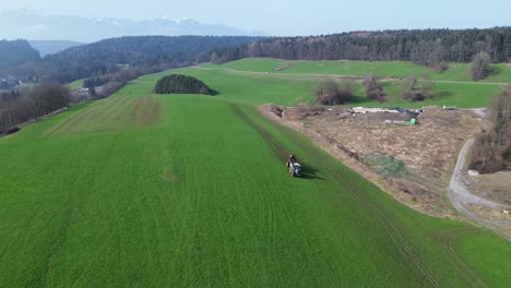 Luftaufnahme-Einer-Drohne,-Die-Einen-Traktor-Mit-Dünger-Auf-Einer-Grünen-Wiese-Mit-Schneebedeckten-Bergen-Im-Hintergrund-Sprüht