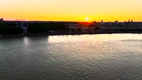 Place-des-Quinconces-Bordeaux-France-Garonne-river-shore-with-Ferris-Wheel-illuminated-during-sunset,-Aerial-approach-shot