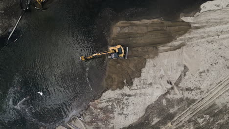 Drone-gets-closer-to-a-yellow-excavator-as-it-cleans-up-a-river-and-piles-dirt-on-shore