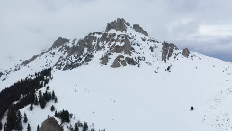 Schneebedeckte-Ciucas-Berge-Unter-Bewölktem-Himmel,-Kiefern-Im-Vordergrund,-Luftaufnahme