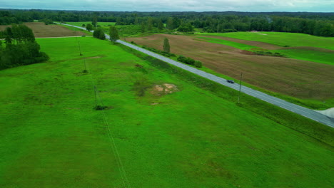 Aerial-drone-landscape-in-wet-green-dead-grass-road-through-countryside-fields-agricultural,-car-drives-slowly-with-humid-daylight-blue-skyline,-outskirts-village