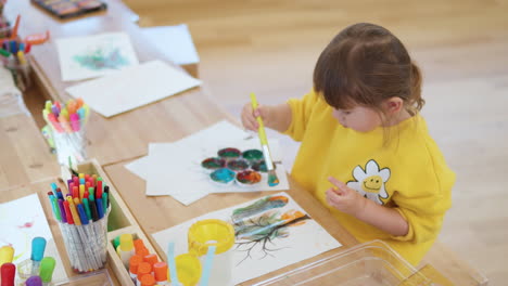 Cute-child-girl-kid-sitting-bythe-desk-using-pintbrush-drawing-and-painting-on-the-paper
