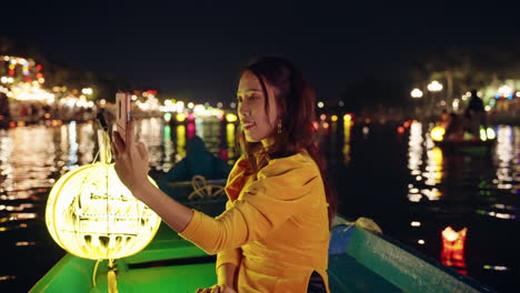 Una-Bonita-Mujer-Vietnamita-Con-Un-Tradicional-Vestido-Amarillo-Toma-Fotos-Con-El-Teléfono-En-Un-Barco-Nocturno-En-Hoi-An,-Vietnam