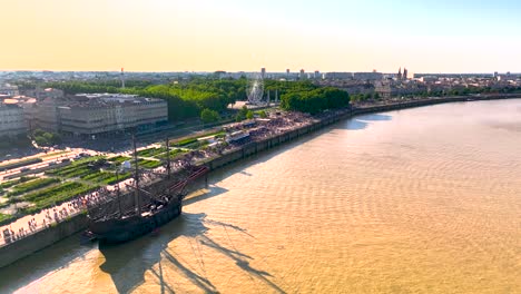 Galleon-sailing-ship-docked-in-the-Garonne-river-shore-during-yearly-Wine-Fair-with-crowds-nearby,-Aerial-flyover-shot