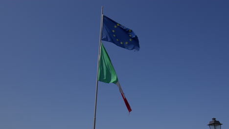 Las-Banderas-De-Italia-Y-Europa-Ondeando-Una-Al-Lado-De-La-Otra-Contra-Un-Fondo-De-Cielo-Azul-En-Bellagio,-Lombardía,-Italia---Tiro-De-ángulo-Bajo