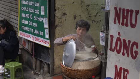 Woman-eating-street-food-in-Hanoi,-steam-rising-from-large-pot-on-a-busy-sidewalk,-daytime