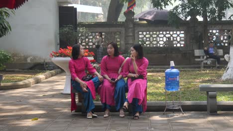 Three-Vietnamese-women-in-Ao-Dai-dresses-talking-on-a-bench,-Hanoi