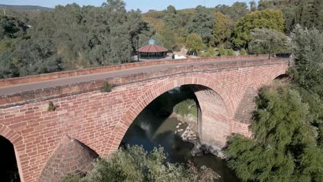 Aérea-Del-Puente-Del-Siglo-XVI-Sobre-El-Río-Guadalquivir,-Cruzando-Edificios-Y-Jardines-Del-Balneario-De-Marmolejo,-En-La-Provincia-De-Jaén.