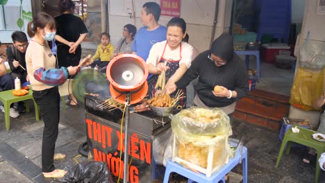 Vibrante-Puesto-De-Comida-Callejera-En-Hanoi-Que-Vende-Deliciosas-Brochetas-De-Carne-A-La-Parrilla-Y-Un-Ambiente-Animado