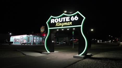 Route-66-large-sign-at-night-in-Kingman,-Arizona-with-vehicles-driving-by-and-stable-video-at-an-angle