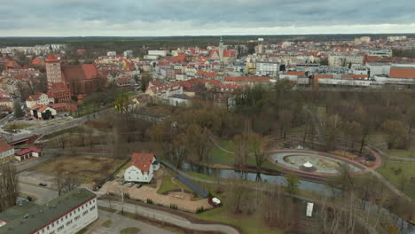 aerial-view-of-Olsztyn,-Poland,-clusters-of-traditional-and-modern-architecture,-red-tiled-roofs,-historical-structures,-winding-river-Łyna,-backdrop-of-the-city's-greenery-and-overcast-skies