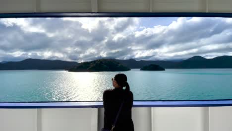 Single-woman-looks-at-beautiful-view-of-water,-islands,-and-cloudy-sky-framed-by-boat-railing-and-ceiling-in-New-Zealand