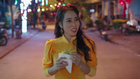 Slow-motion-follow-shot-of-young-Vietnamese-woman-in-traditional-outfit-eating-local-pizza-at-night-in-Hoi-An,-Vietnam
