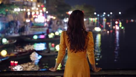 Mittlere-Aufnahme-Von-Hinten-Einer-Frau-Im-Vietnamesischen-Gelben-Kleid,-Die-Die-Nächtlichen-Spiegelungen-Des-Flusses-In-Hoi-An-Betrachtet