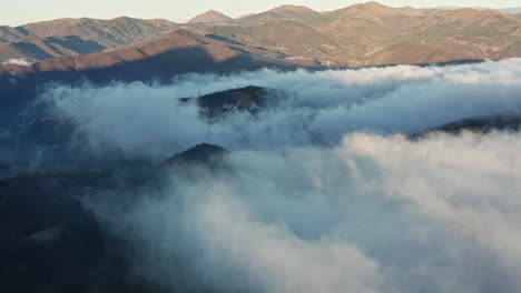 Misty-mountains-with-clouds-rolling-over-the-peaks-at-sunrise,-aerial-view