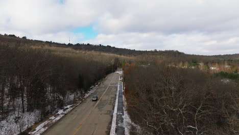 Toma-Aérea-De-Una-Carretera-Con-Un-Coche-Pasando-Por-Un-Bosque-Invernal-Sin-Hojas,-Arkansas