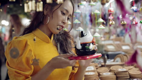 Mujer-Vestida-De-Amarillo-Ao-Dai-Admirando-Un-Recuerdo-De-Gato-Afortunado-En-El-Mercado-De-Hoi-An,-Colores-Vibrantes,-Noche