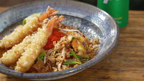 Beautifully-plated-traditional-pad-thai-topped-with-three-fried-shrimp-tempura-sticks-on-blue-stoneware-dish,-close-up-slider-4K