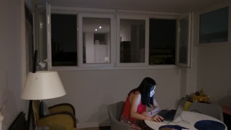 A-woman-is-using-a-laptop-indoors-while-a-thunderstorm-is-taking-place-outside,-with-flashes-visible-through-the-home-windows