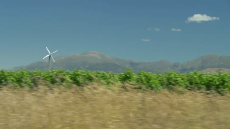 Driving-by-a-vineyard-with-a-small-windmill-and-mountains-in-background-in-slow-motion---Marlborough-Wine-District,-New-Zealand