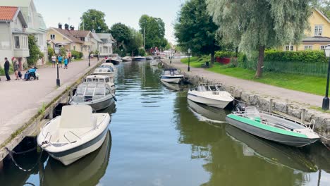 Boats-docked-by-canal-in-small-town-in-summertime-Sweden,-people-walk