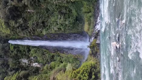 Vertikale-Eines-Wasserfalls-Namens-Thunder-Creek-Falls,-Der-In-Einen-Fluss-Fließt,-Der-Von-üppigem-Regenwald-An-Der-Westküste-Neuseelands-Umgeben-Ist