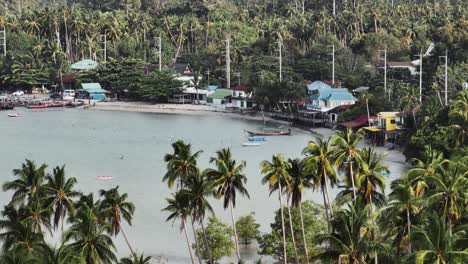 Drone-footage,-flying-over-palm-trees-and-bay-with-houses-and-huts