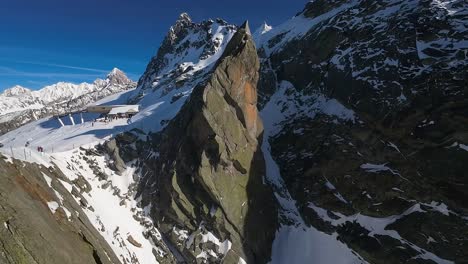 Soaring-aerial-shot-of-a-sharp-mountain-precipice-against-the-backdrop-of-Chamonix-Mont-Blanc's-ski-slopes-on-a-sunny-winter-day