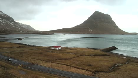 Stunning-4K-drone-footage-captures-the-aerial-beauty-of-a-picturesque-lake,-featuring-a-charming-red-roofed-house-and-a-car-traveling-along-the-nearby-road-in-a-cinematic-and-distinctive-manner