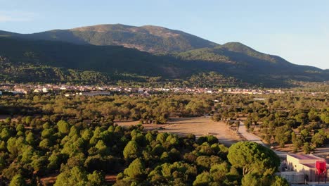 Descent-flight-with-in-the-golden-hour-of-the-afternoon-over-green-pine-forest-with-yellow-summer-meadows-with-background-of-forested-mountains-with-a-sunny-village-slopes-and-blue-sky-in-Avila-Spain