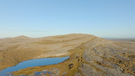 Drone-slowly-ascends-above-curved-slope-hillside-with-patches-of-grass-under-blue-sky,-the-Burren-Ireland