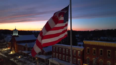 American-flag-in-front-of-winter-sunset-over-a-small-town-in-USA-covered-in-snow