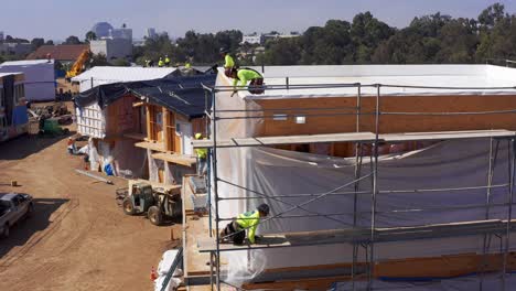 Close-up-panning-aerial-shot-of-a-construction-crew-working-on-the-siding-of-a-modular-housing-unit-at-a-building-site-in-West-Los-Angeles,-California