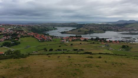 flight-with-a-drone-at-the-mouth-of-a-river-to-the-marcon-a-turn-of-the-camera-we-see-a-hill-with-horses-grazing-and-we-see-a-town-with-green-meadows-on-a-cloudy-morning-in-Cantabria-Spain