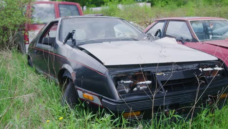 1980s-Mustang-fox-body-without-headlights,-sitting-abandoned-next-to-other-vintage-cars-in-the-grass