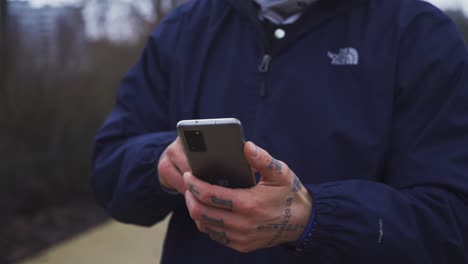 Male-outdoor-fitness-instructor-using-smartphone-in-rain-coat,-closeup-of-tattooed-hands-touching-smartphone-for-online-workout