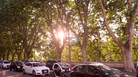 Toma-Panorámica-En-Cámara-Lenta-De-Destellos-De-Lentes-Mientras-El-Sol-Se-Asoma-A-Través-De-Hermosos-árboles-En-La-Carretera-De-La-Ciudad-Italiana-Con-Tráfico