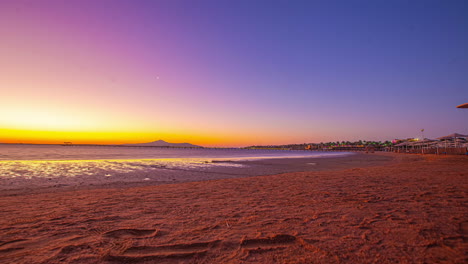 Time-lapses-of-the-sea-on-the-beach-with-the-orange-sky,-small-buildings-and-palm-trees-in-the-background