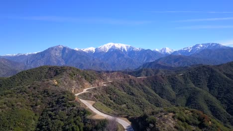 A-reverse-aerial-flyover-of-a-winding-mountain-road,-heading-towards-snowcapped-mountians-in-the-background