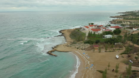 Aerial-view-of-a-coastal-area-blending-natural-and-urban-landscapes,-with-rocky-terrain-leading-to-agricultural-fields,-and-a-scattering-of-buildings-with-a-town-in-the-distance---Paralimni,-Cyprus