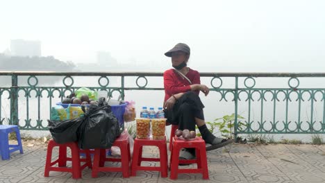 Street-vendor-seated-by-Tay-Ho-Lake,-Hanoi,-selling-snacks-with-misty-backdrop