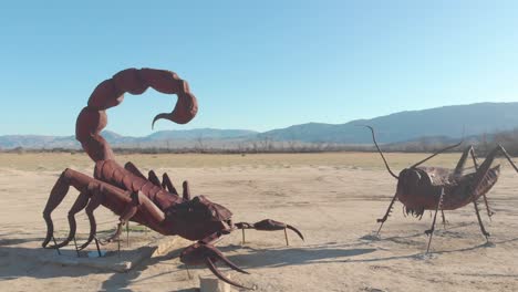 semi-orbit-drone-shot-of-2-statues-of-a-scorpion-and-a-grasshopper-facing-each-other-in-the-desert-with-mountains-in-the-background