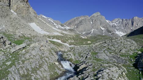 Mountain-stream-flowing-through-rocky-terrain-with-peaks,-Cascata-di-Stroppia-and-Lago-Niera-in-the-background,-sunny-day
