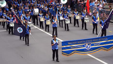 Students-from-Mackay-North-State-High-School-Marching-Band-performing-and-walking-down-the-Street-during-the-Anzac-Day-parade,-with-crowds-gathered-alongside-to-honour-the-memory-of-those-who-served