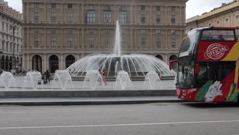 Handheld-clip-of-red-tour-bus-in-front-of-famous-landmark-fountains-in-Piazza-De-Ferrari-in-Genoa,-Italy